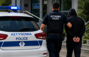 Αστυνομικός συνοδεύει πρόσωπο με χειροπέδες δίπλα σε περιπολικό